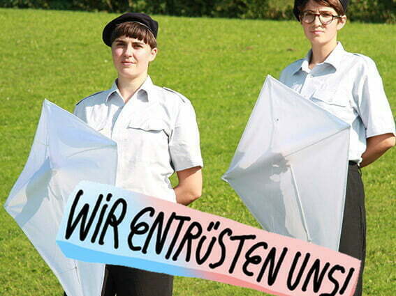 Zwei Frauen stehen mit einem Plakat mit dem Text: «Wir entrüsten uns!» auf einer Wiese.