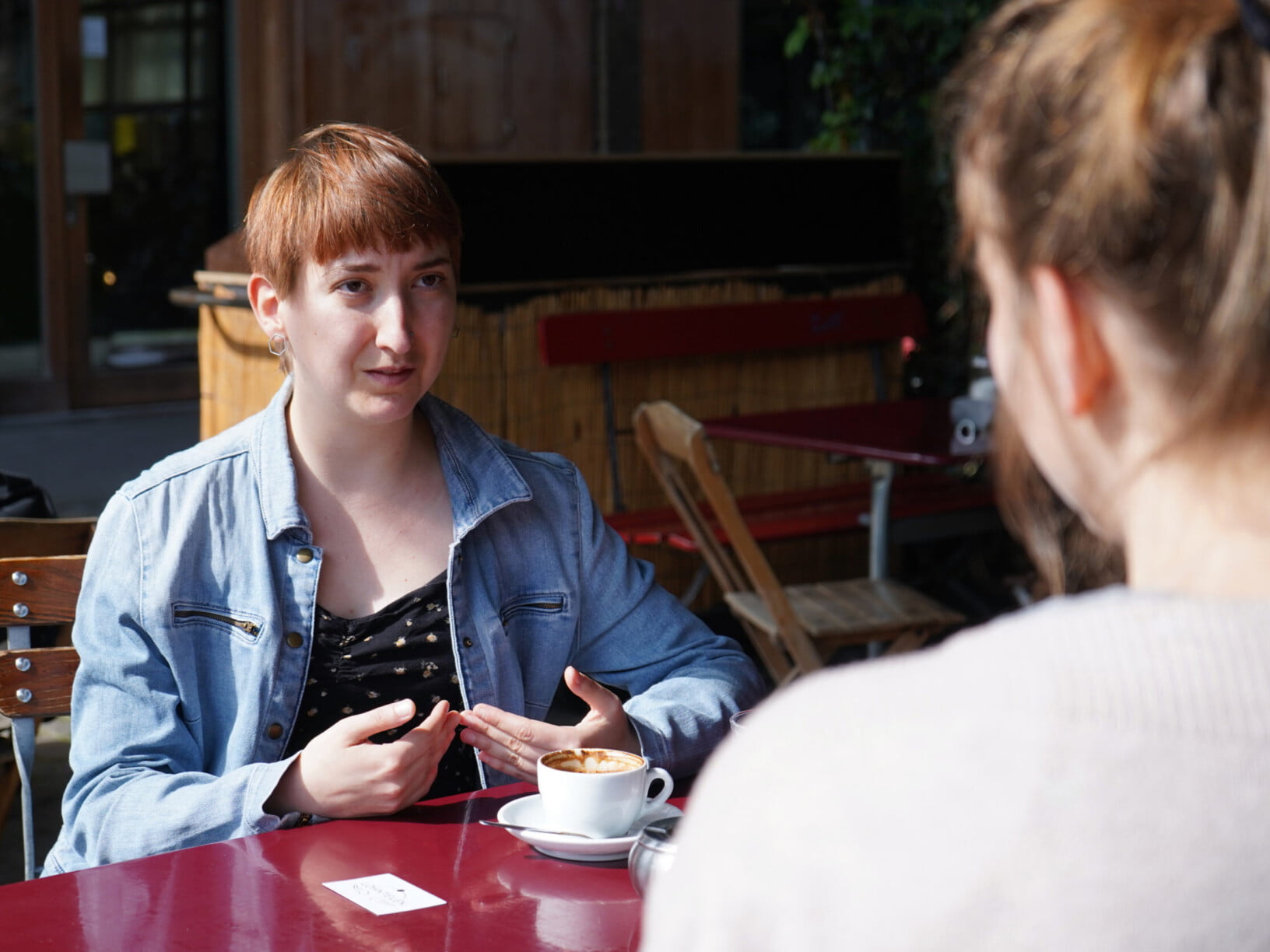 Eine Frau mit kurzen Haaren spricht mit einer anderen Frau. Sie trägt eine blaue Jacke und trinkt einen Kaffee. Sie sitzen an einem roten Tisch an der Sonne..