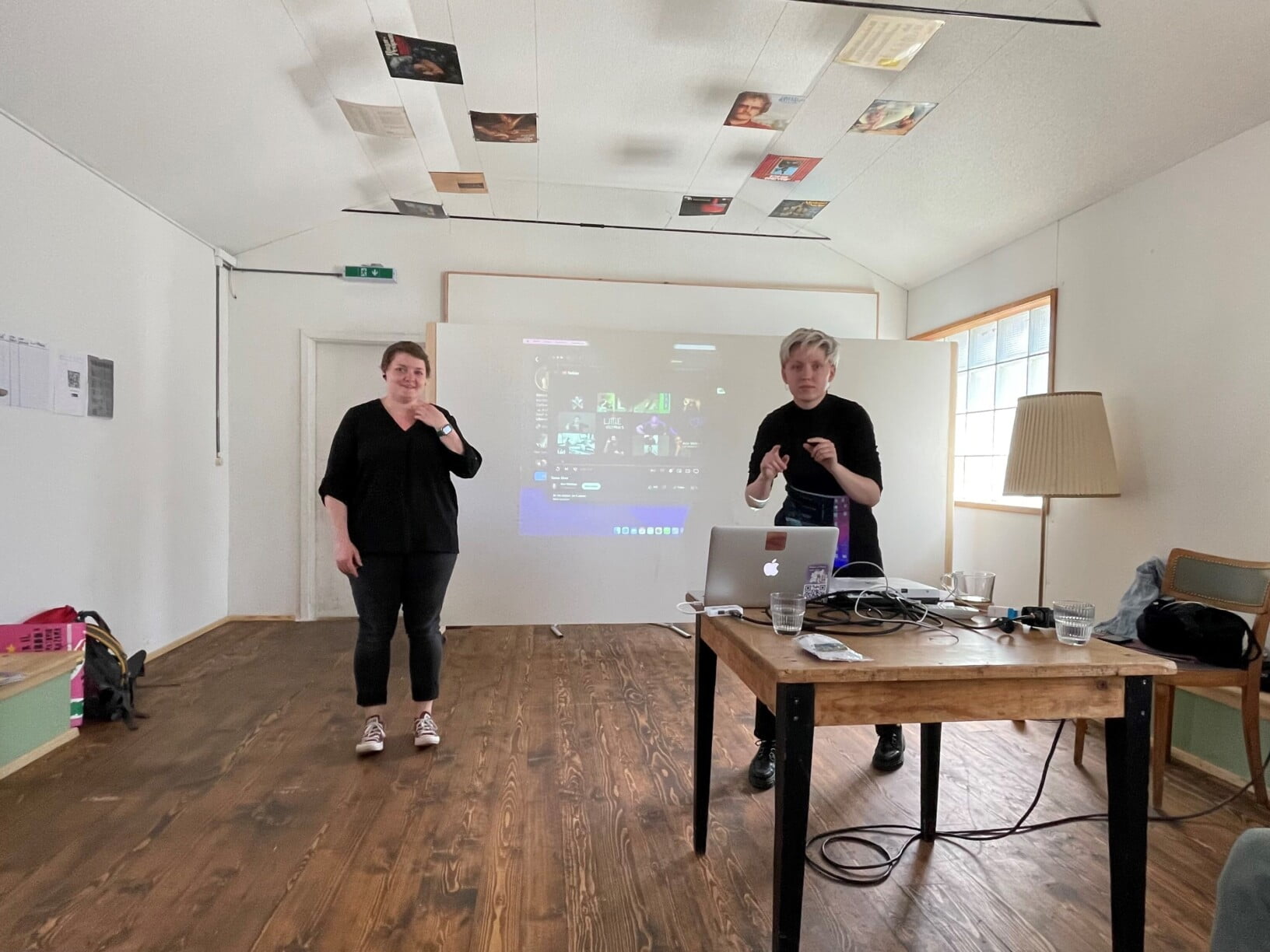 Zwei Personen stehen vor einer weissen Leinwand, sie schauen nach vorne. Der Boden ist aus Holz, vor ihnen steht ein Tisch mit einem Laptop.