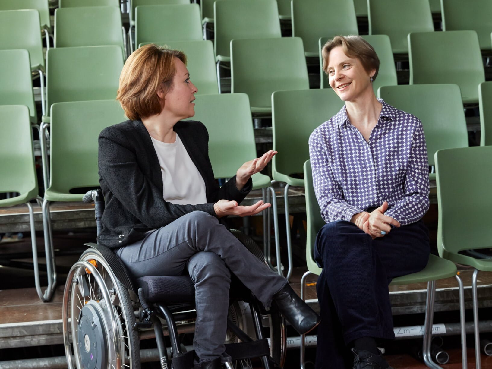 Zwei Frauen sprechen miteinander, im Hintergrund gibt es grüne Stühle, die Frau links sitzt in einem Rollstuhl, die andere auf einem grünem Stuhl. Sie lächeln.