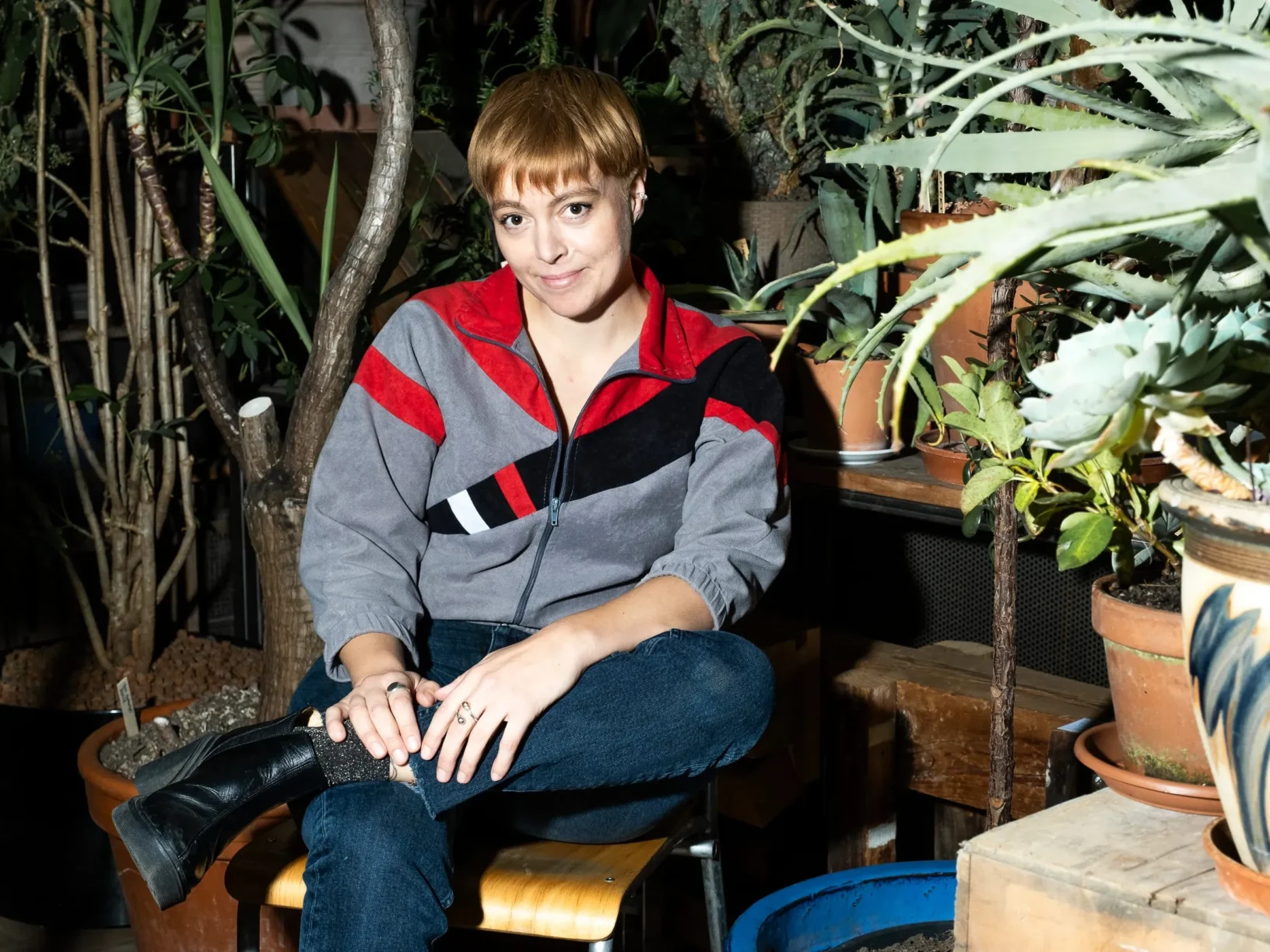 Silja Gruner sitzt auf einem Holzstuhl, den linken Fuss über ihr rechtes Knie gelegt und lächelt in die Kamera. Um sie herum stehen viele grosse Pflanzen. Die eigentlich dunkle Szenerie wird erst durch den Kamerablitz erleuchtet. Bildquelle: Nathi Jufer
